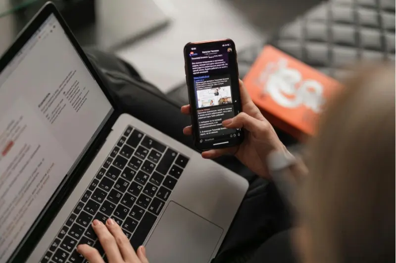 A woman reading the news on her phone and using a laptop